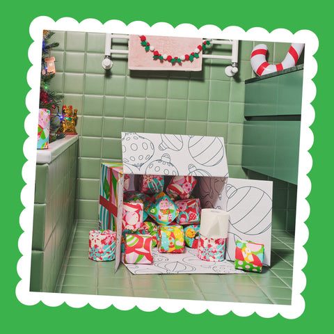 An open Gift Edition box full of colourfully wrapped toilet paper rolls on the floor of a festive green-tiled bathroom.