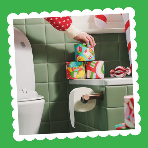 A hand places a wrapped Gift Edition roll on a stack of festive rolls on a bathroom shelf above a toilet roll holder.